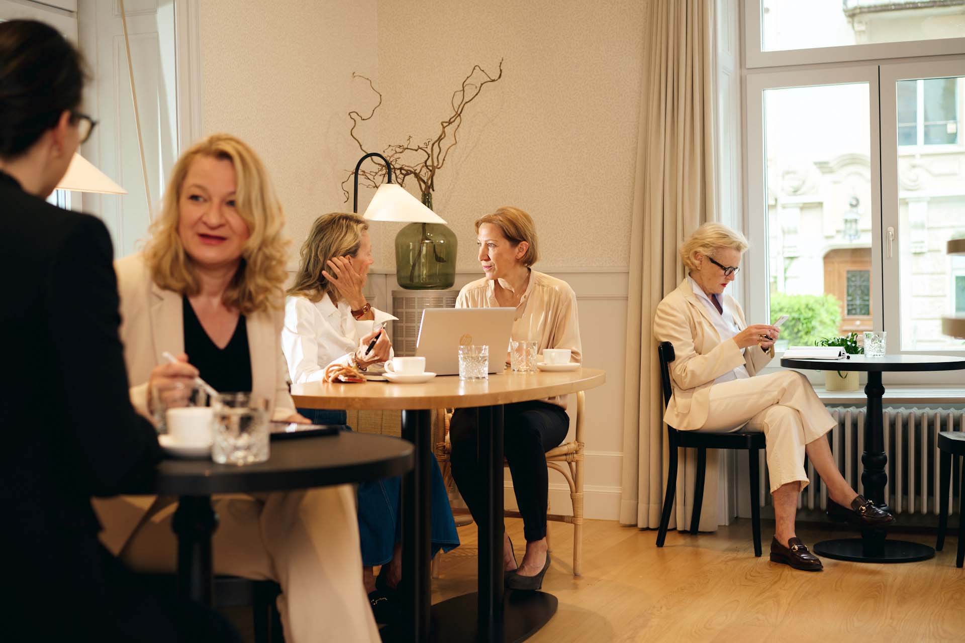  Fünf Frauen im Business Look sitzen im schlicht aber elegant eingerichteten Bistro des Hotel Alma im Seefeld. An zwei Tischen führen je zwei Frauen Gespräche. Eine dritte Frau sitzt an einem kleinen Tisch im Hintergrund und schaut auf ihr Mobil.