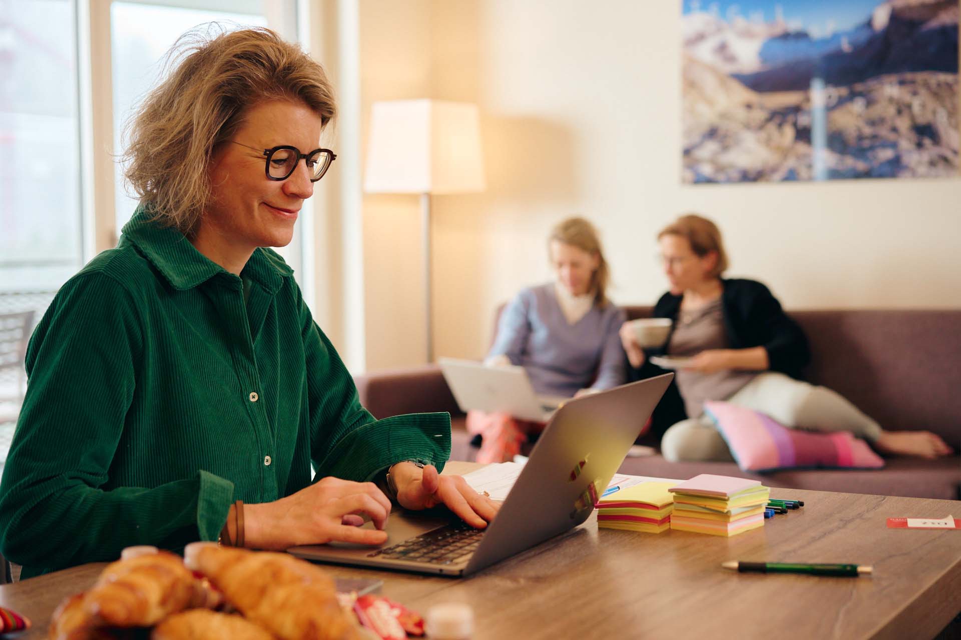 Nicoletta in dunkelgrün gekleidet sitzt in ihren Laptop schauend am Esstisch im Peaks Place. Im Hintergrund sitzen Karin (links) und Isabelle (rechts) auf dem Sofa und schauen in Karins Laptop.