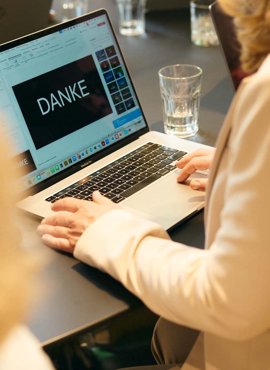 Eine in beige gekleidete Frau arbeitet sitzend auf ihrem Laptop an einer Präsentation. Ebenfalls auf dem Tisch steht ein fast leeres Wasserglas.