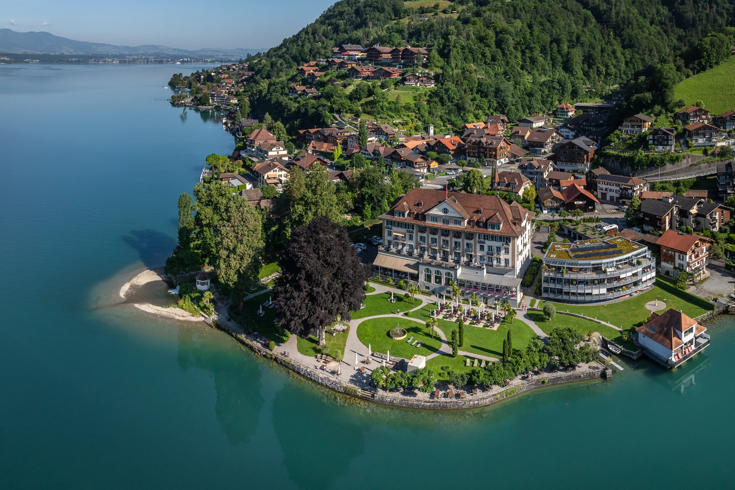 Luftaufnahme des Parkhotels Gunten am Thunersee mit Park, Seeufer und dem Dorf Gunten am bewaldeten Hang